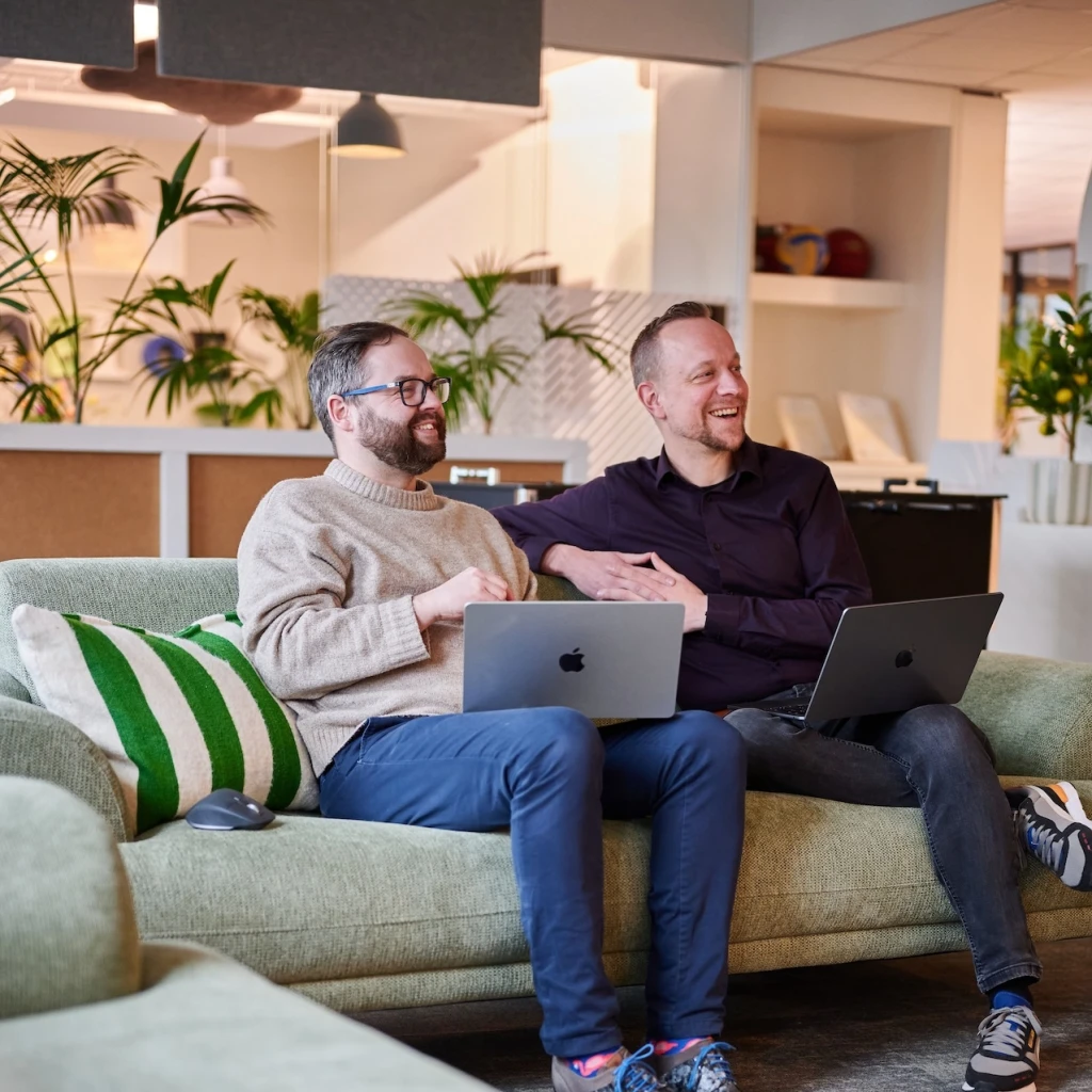 Twee mensen zitten op een bank met laptops in een gezellige ruimte.